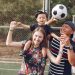 Positive smiling diverse parents giving piggyback ride to adorable toddler son while spending weekend together with football ball on sports ground on sunny day